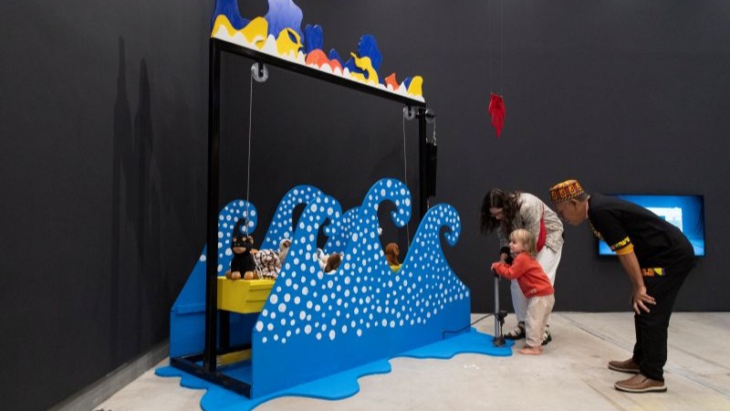 Criança pequena interage com instalação enquanto dois adultos a acompanham. A obra é composta por um painel com moldura preta e formas onduladas azuis com bolinhas brancas, lembrando ondas do mar. Dentro da instalação há brinquedos de pelúcia.