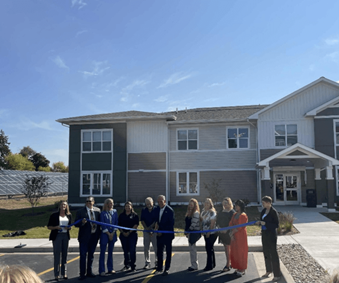 A group of people standing outside of a residential building holding a long blue ribbon. A group of people standing outside of a residential building holding a long blue ribbon.