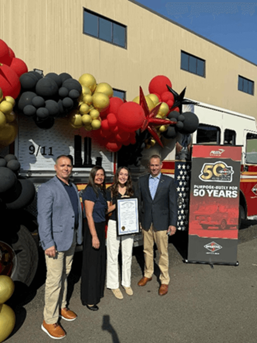 Staff members presenting a tribute certificate to members of the Spartan Fire Chassis in front of a large balloon display and a banner that reads "Purpose Built For 50 Years" with a picture of a firetruck on it. Staff members presenting a tribute certificate to members of the Spartan Fire Chassis in front of a large balloon display and a banner that reads "Purpose Built For 50 Years" with a picture of a firetruck on it.