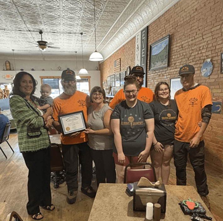 Michigan state Representative Angela Witwer holding a baby while presenting staff at the Big Tree Worx LLC with a tribute certificate. Michigan state Representative Angela Witwer holding a baby while presenting staff at the Big Tree Worx LLC with a tribute certificate.