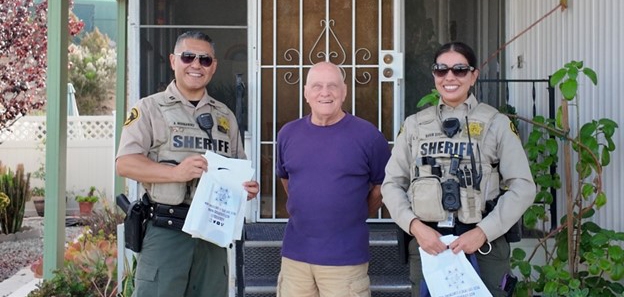 Two male deputies wearing vest with bags taking a photo with an elderly man wearing a purple shirt outside of his house.