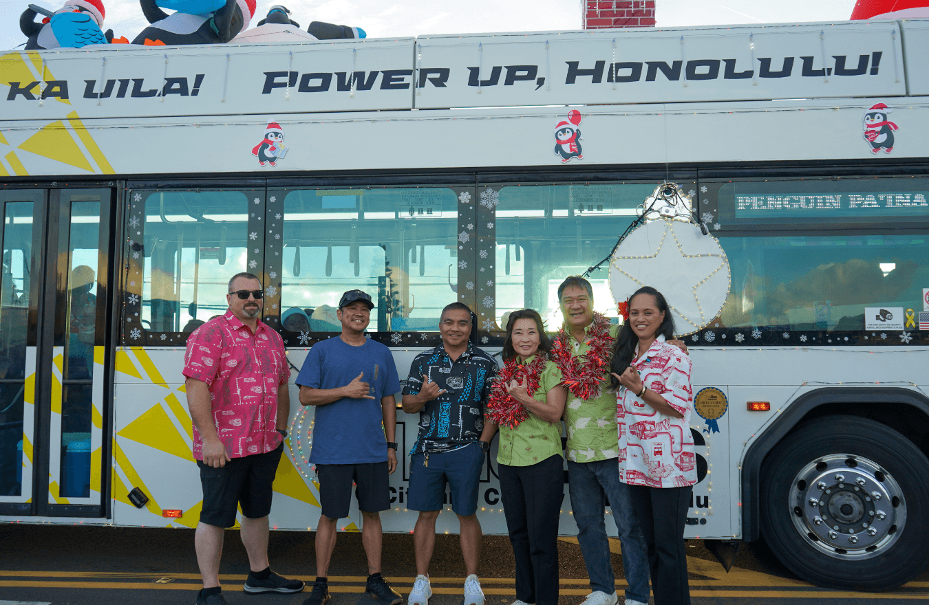 Lt. Gov. Sylvia Luke and group smiling while holding up shakas for a group photo in front of a bus with Christmas decorations. 
