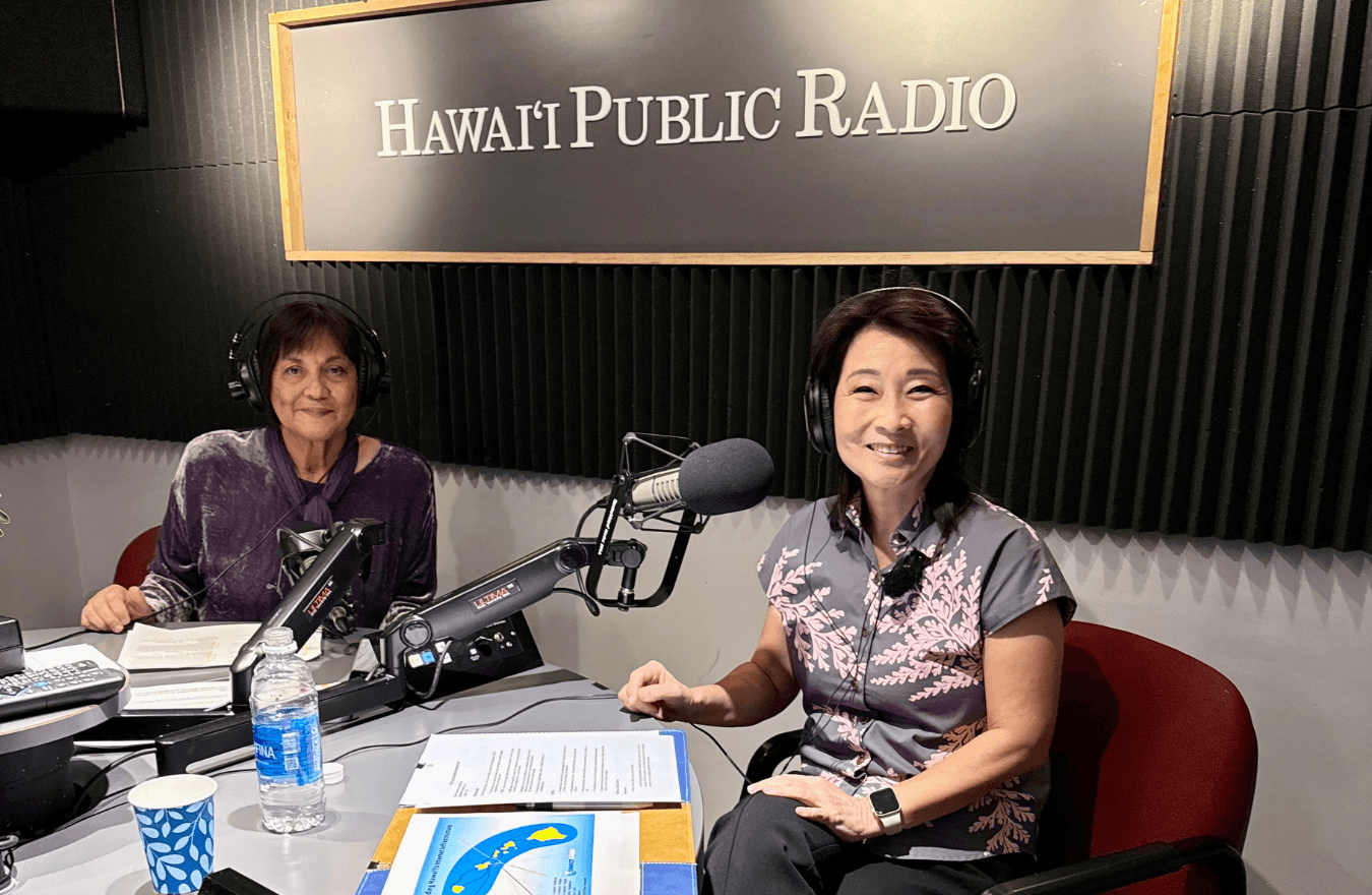 Lt. Gov. Sylvia Luke and a lady smiling while sitting at a table in front of microphones with headphones on in front of a sign saying, “Hawaiʻi Public Radio.”  
