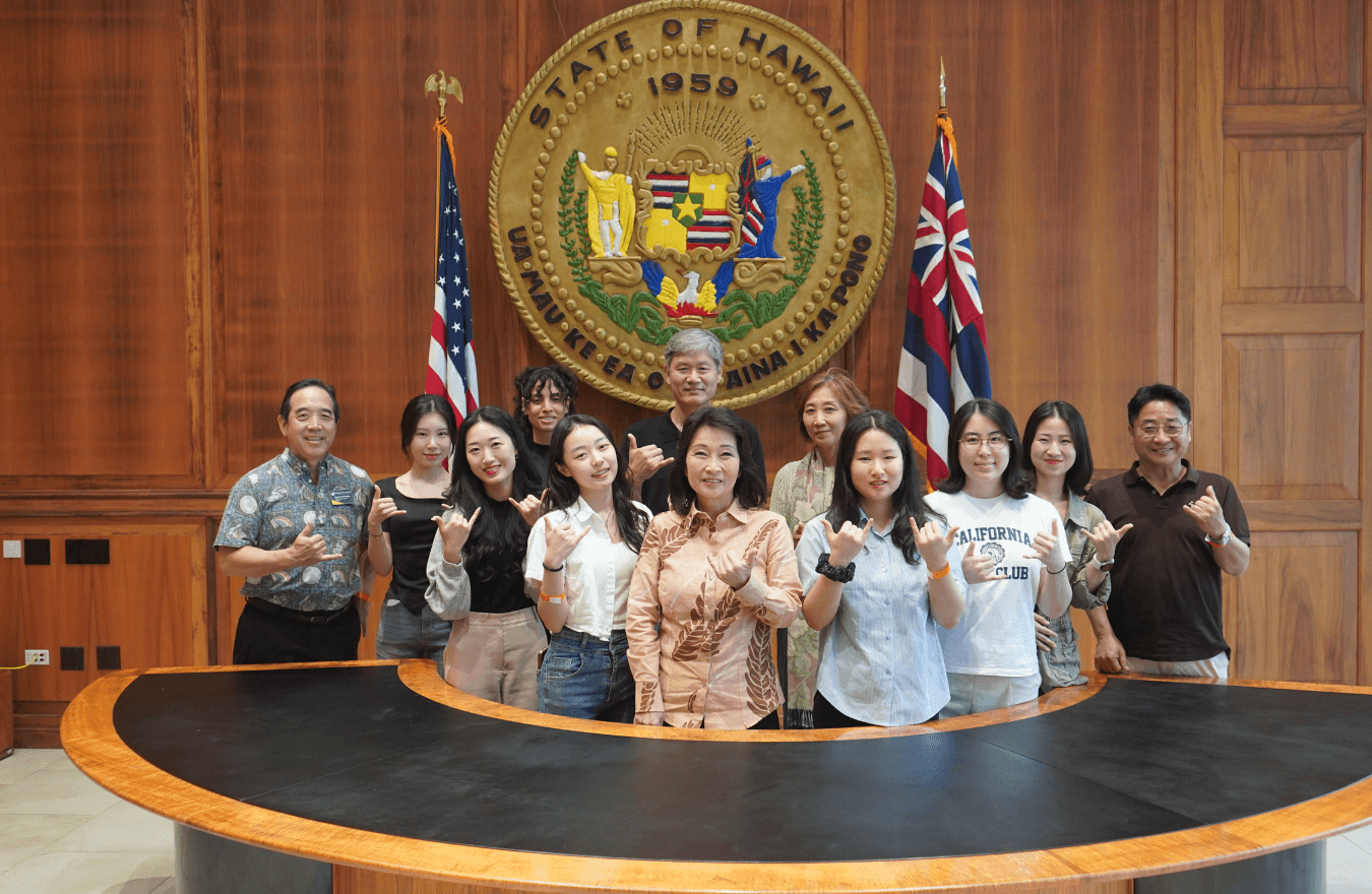 Lt. Gov. Sylvia Luke and group smiling and holding up shakas for a group photo in front of the Hawaiʻi State Seal. 