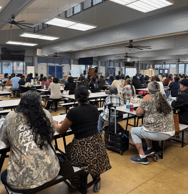 Large group of people sitting at tables faced toward a stage in a cafeteria. 