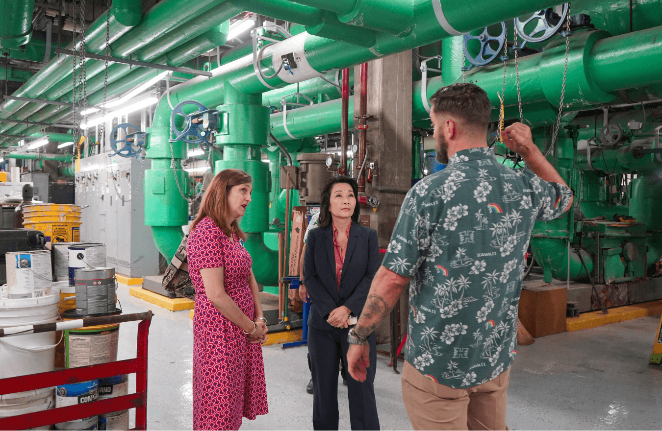 Lt. Gov. Sylvia Luke and a lady looking at a man speaking while standing in a room with big, green pipes. 