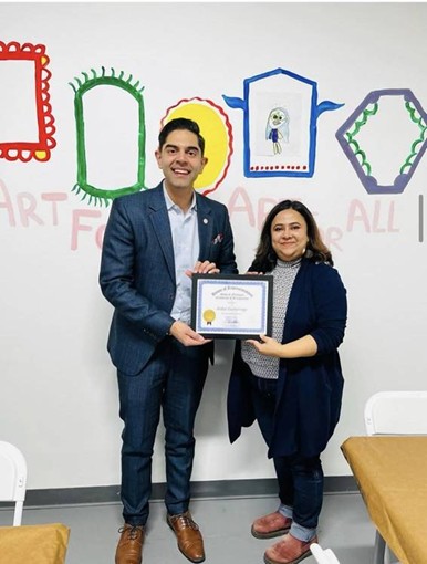 Democratic Leader Ranjeev Puri with the owner of Artful Gatherings holding their framed certificate for February Business of the Month in front of a wall with picture frames painted on it. Democratic Leader Ranjeev Puri with the owner of Artful Gatherings holding their framed certificate for February Business of the Month in front of a wall with picture frames painted on it.