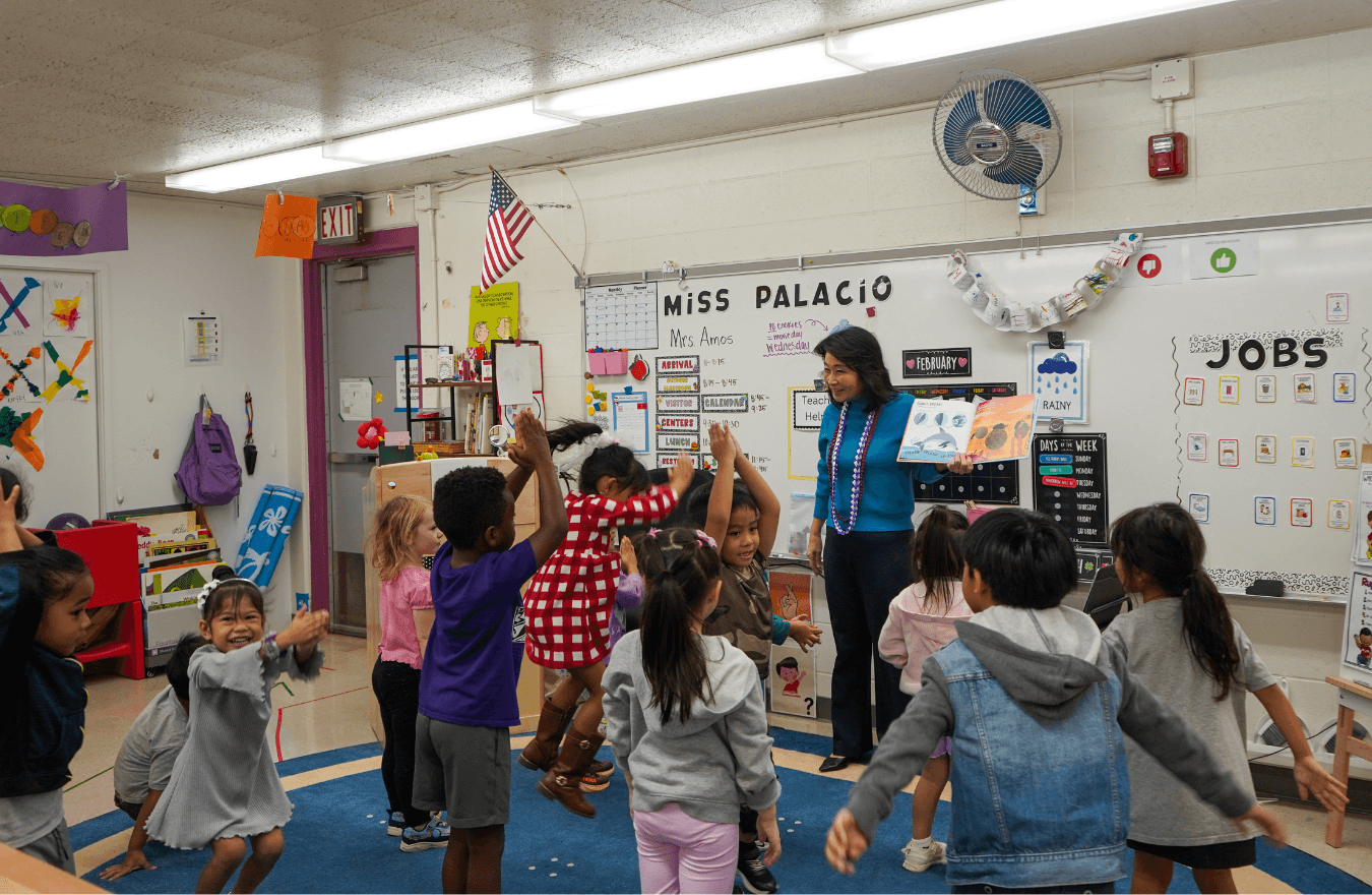 Lt. Gov. Sylvia Luke standing up and smiling while holding a book open and looking at standing preschool students. 
