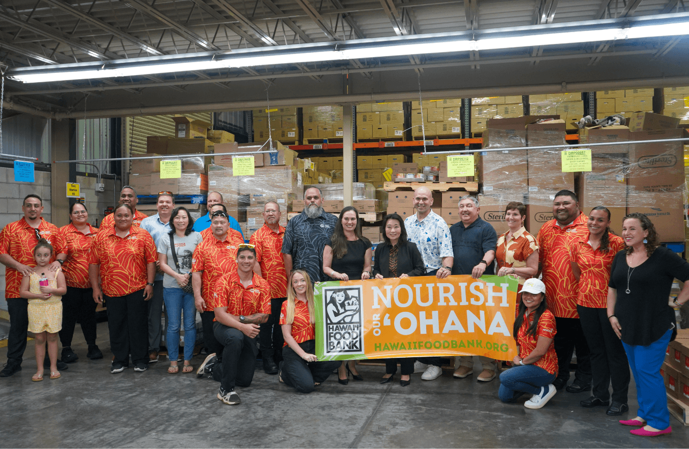 Lt. Gov. Sylvia Luke and group standing in a warehouse in front of large, stacked boxes, holding a sign that says, “Nourish our ʻOhana.” 