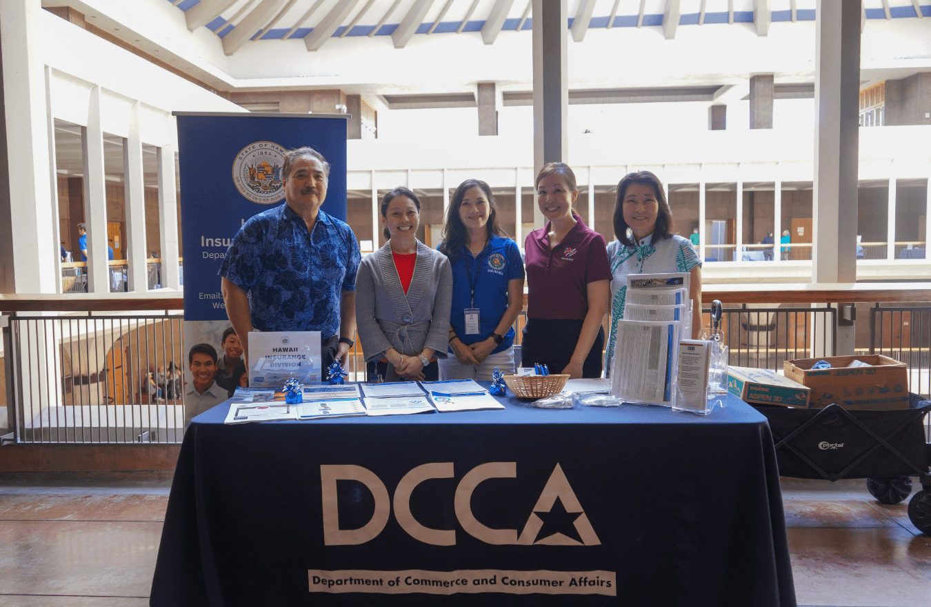 Lt. Gov. Sylvia Luke and group smiling for a group photo in back of a table that says, “DCCA Department of Commerce and Consumer Affairs.” 