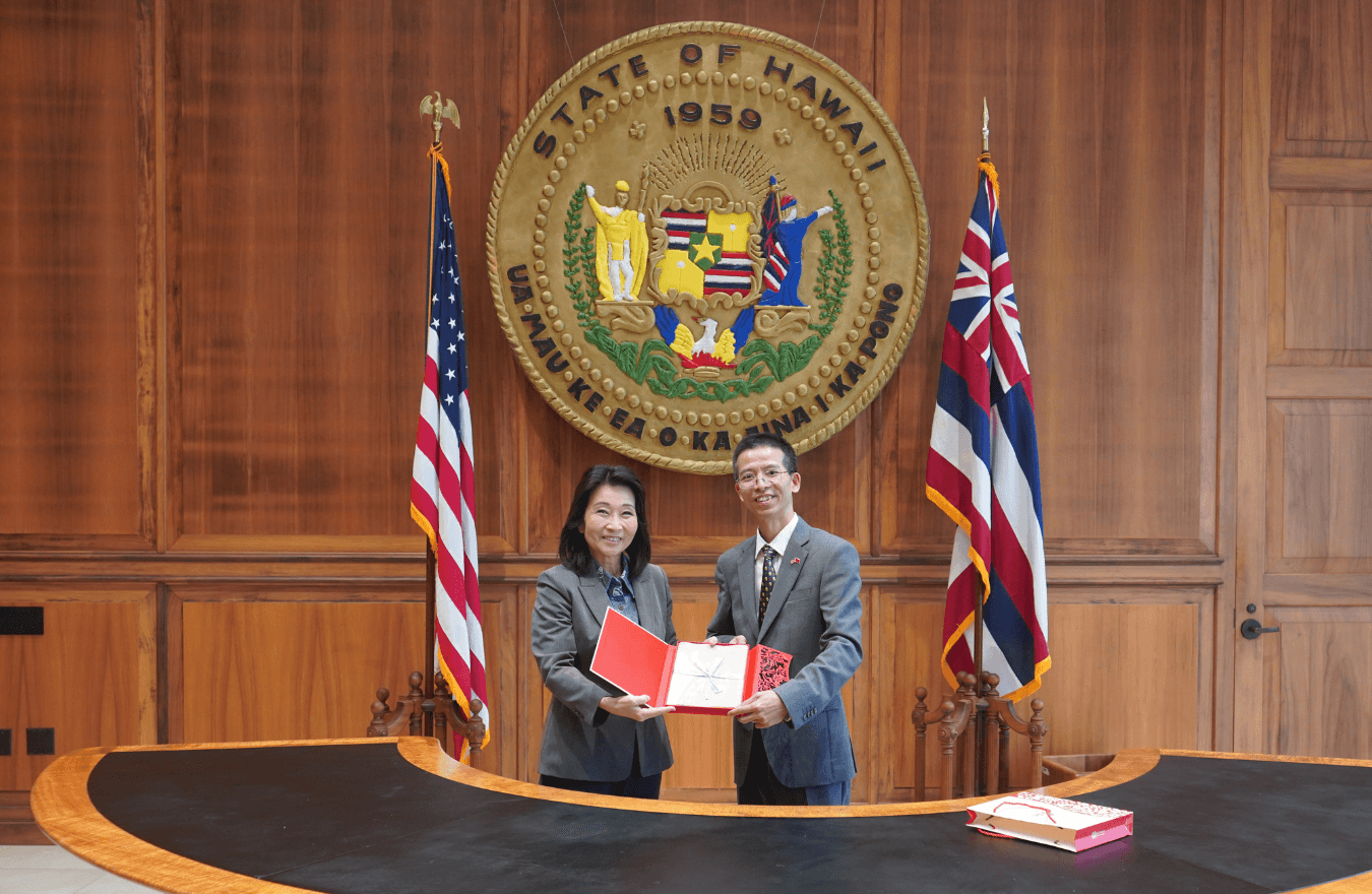 Lt. Gov. Sylvia Luke and man holding open a red box while smiling for a photo in front of the Hawaiʻi State Seal. 