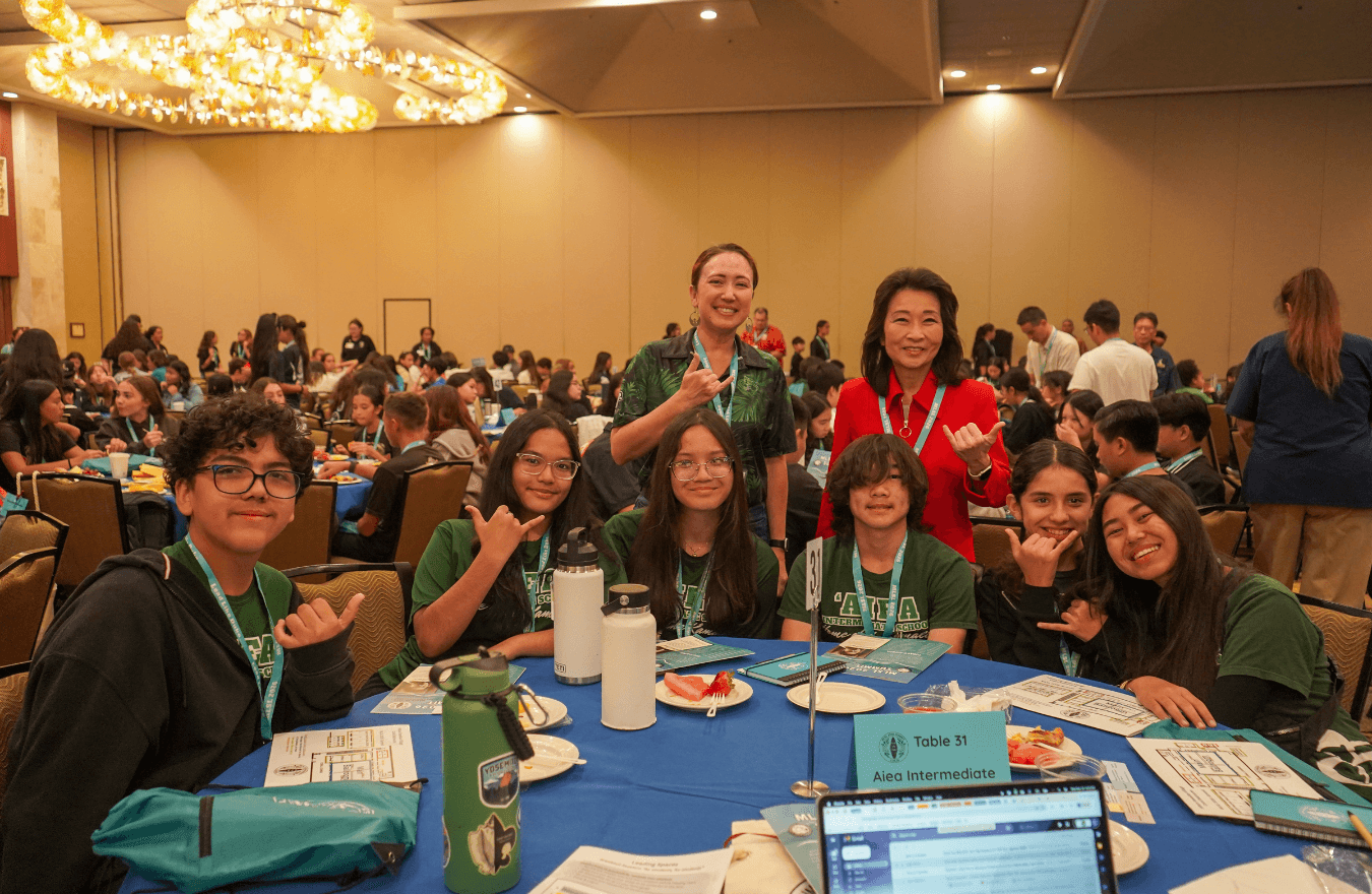 Lt. Gov. Sylvia Luke and group smiling while holding up shakas for a group photo in a banquet hall with lots of tables. 
