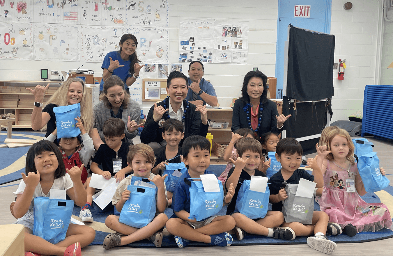 Lt. Gov. Sylvia Luke, a legislator, teachers, and pre-k students holding lunch bags smiling and holding up shakas in a Ready Keiki preschool classroom in Hawaiʻi Kai, Oʻahu. 