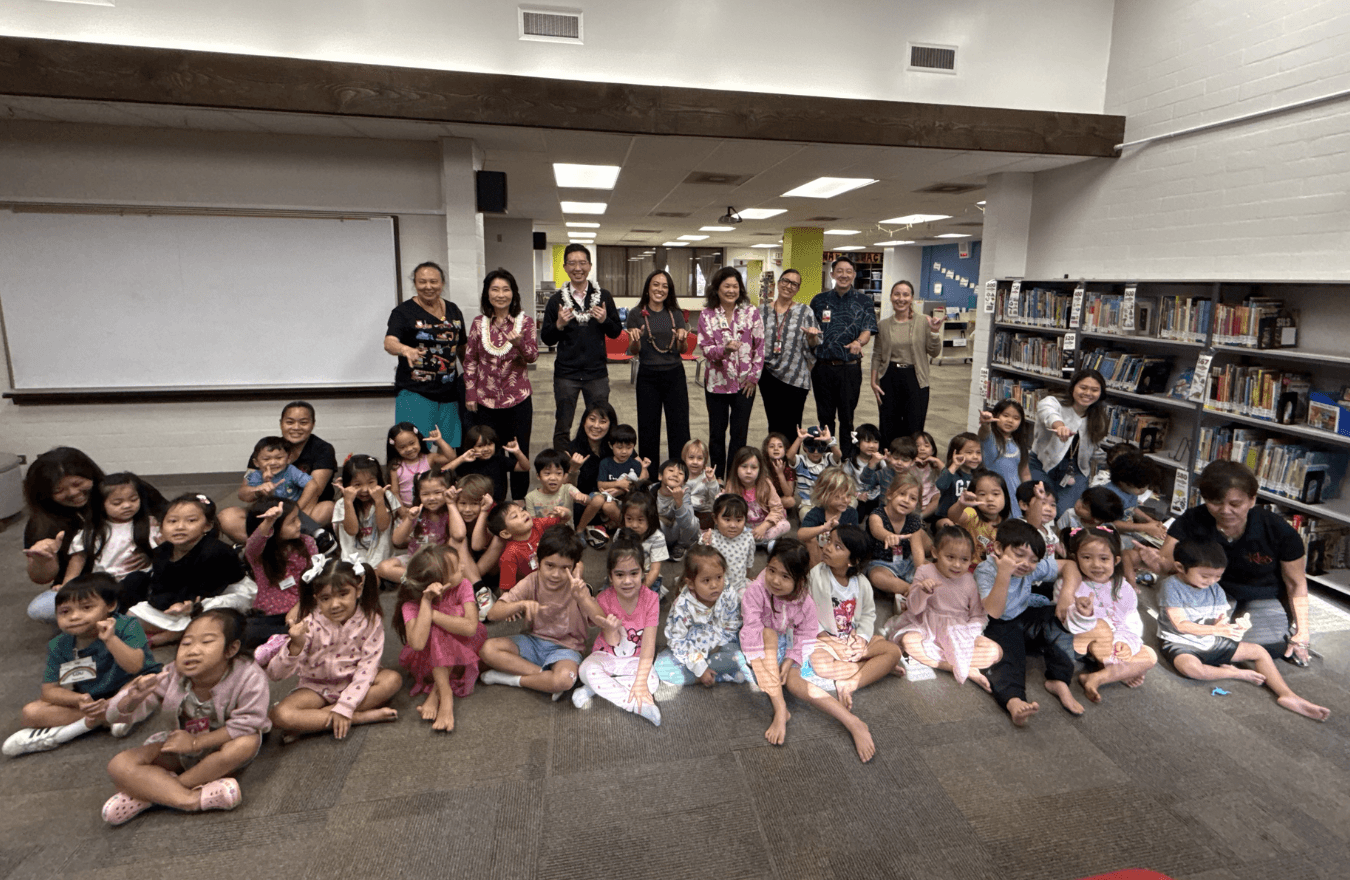 Lt. Gov. Sylvia Luke, legislators, teachers, and pre-k students smiling and holding up shakas in a school library.  