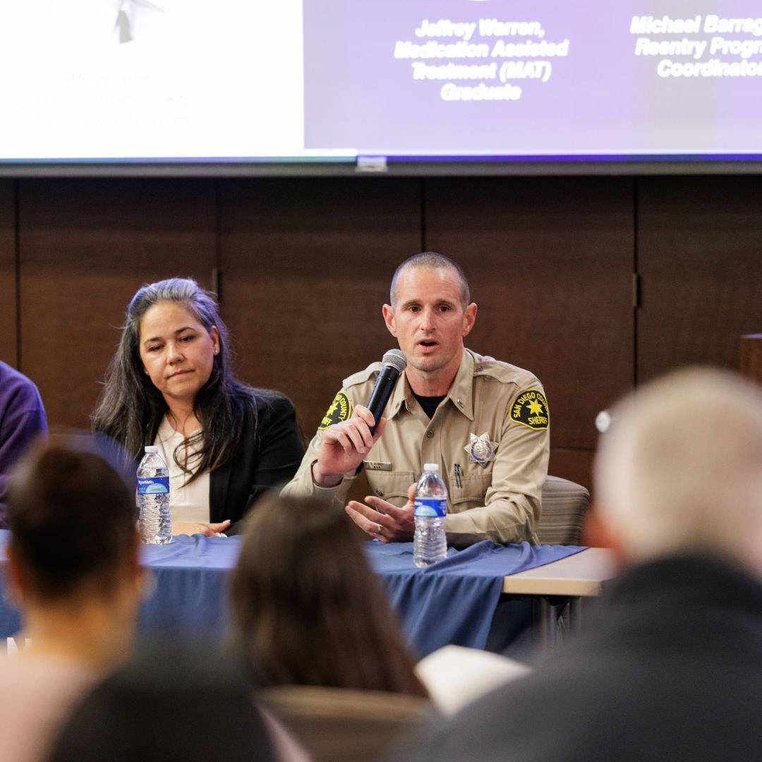 A panel seated at a table during an indoor event. A uniformed individual speaks into a microphone, with water bottles and notes on the table. Other panelists sit beside the speaker, and audience members are visible in the foreground. A presentation screen with text is displayed behind the panel.