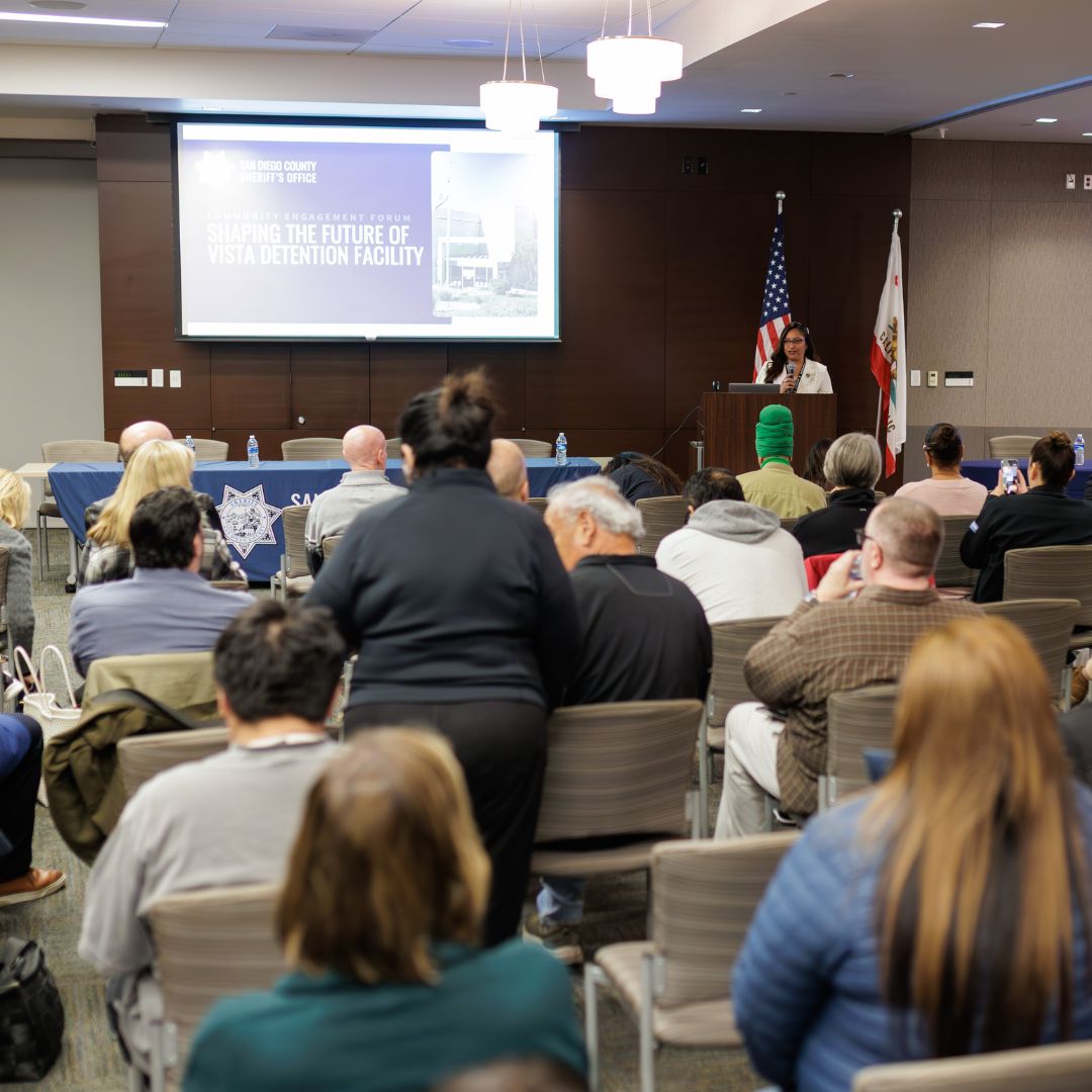 Audience seated in a conference room facing a speaker at a podium. Behind the speaker are U.S. and California flags, and a large projected slide titled “Shaping the Future of Vista Detention Facility.” A long table with a blue tablecloth and bottled water is positioned at the front of the room. Bright ceiling lights illuminate the space.