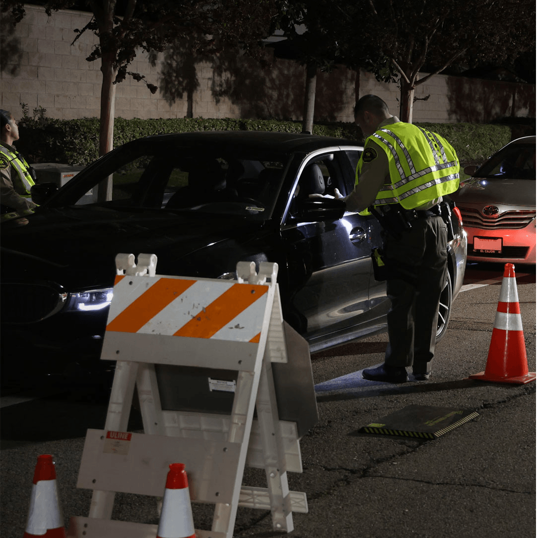 A nighttime roadside safety checkpoint with traffic cones, a reflective road barrier, and a vehicle stopped while a uniformed traffic officer speaks to the driver. Trees and parked vehicles line the background, and the area is lit by street lighting.
