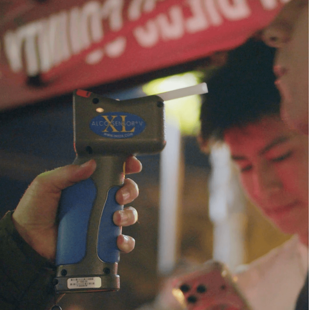 A close-up nighttime scene showing a handheld alcohol-screening device held by a uniformed deputy near an individual who is standing beside others. A red canopy with partially visible text is overhead, and the background includes blurred lights and figures, suggesting an active enforcement area. Let me know if you’d like a shorter or more formal version!