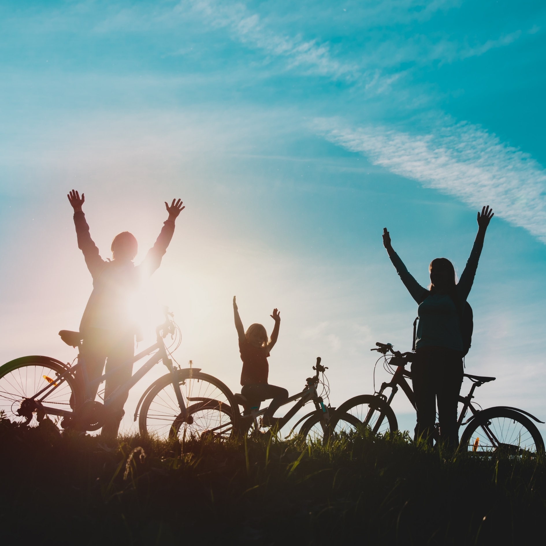 Silhouettes of three individuals with bicycles are seen on a grassy hill at sunset. The bicycles are positioned side‑by‑side, and all three individuals have their arms raised toward the sky. The sun is low on the horizon, creating a bright glow behind the group, while the sky above shows soft clouds and a gradient of blue tones.