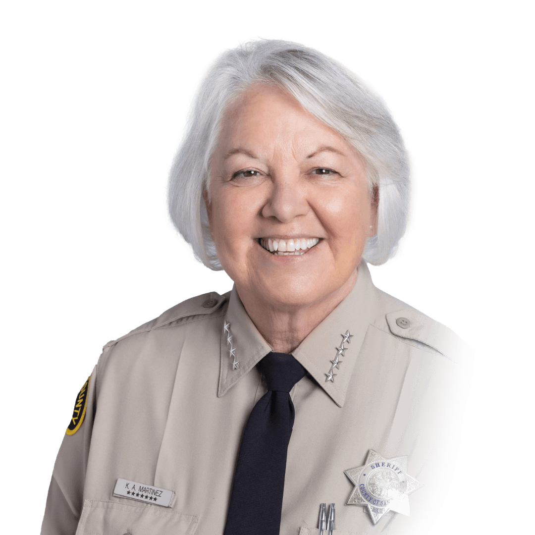Uniformed sheriff’s department member wearing a tan shirt with collar insignia, a dark tie, a nameplate, and a star‑shaped badge.