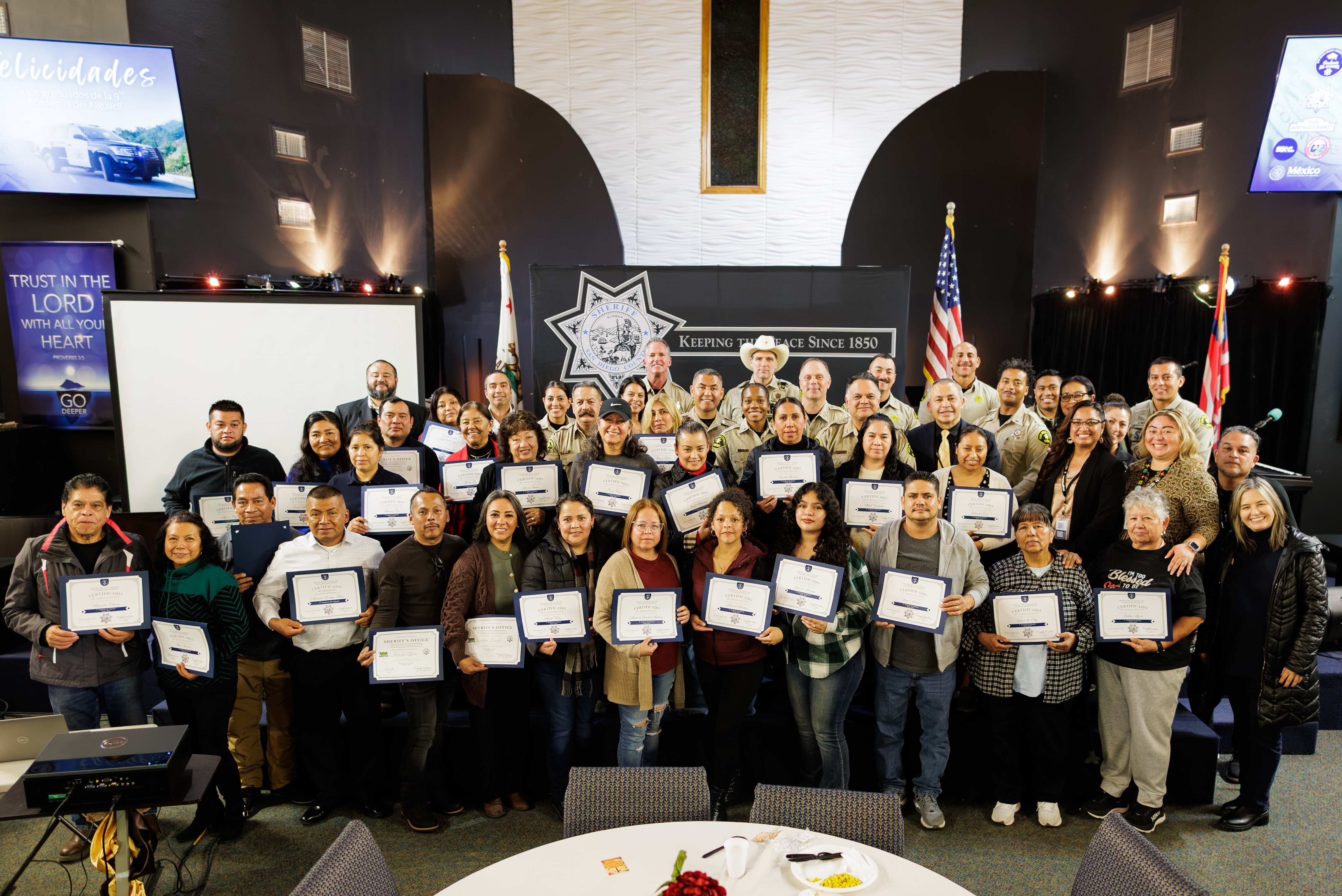 Several individuals stand in a line outdoors holding certificates. A Sheriff’s Department vehicle is parked behind them, displaying a badge emblem on the door. Trees and scattered yellow petals are visible around the paved area.