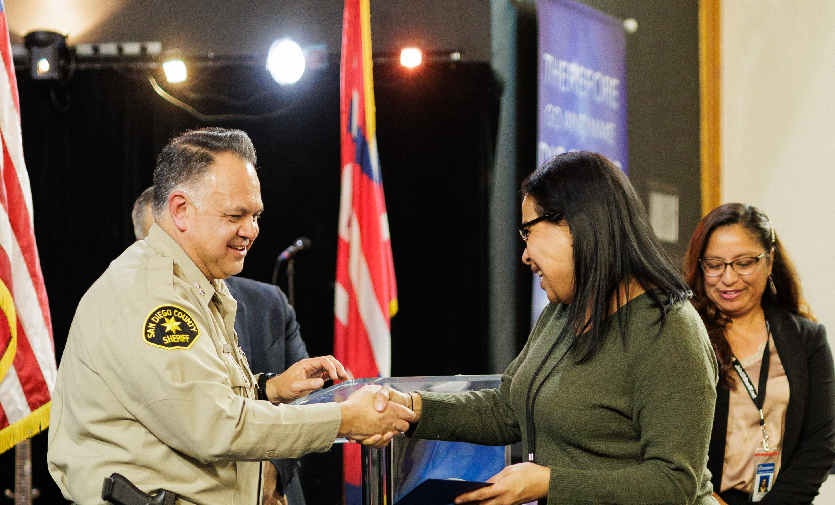 An individual in a San Diego County Sheriff uniform is shaking hands with another individual across a podium during a formal indoor event. A flag stands in the background, along with a banner and stage lighting. Another person stands slightly behind them, observing the interaction.