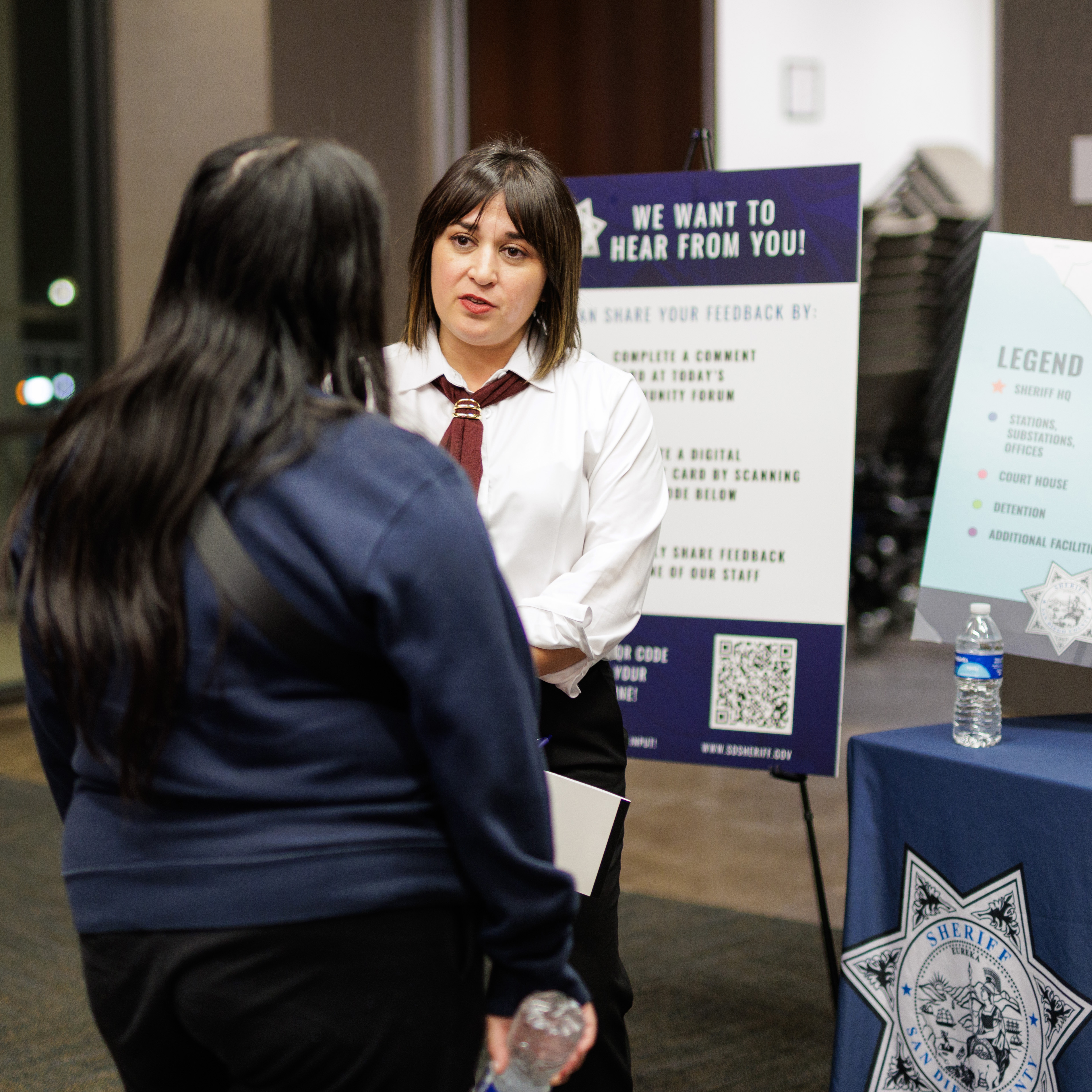 Two individuals are standing and talking near an informational display table. One person is wearing a white shirt with a tie and holding papers, while another person stands facing them holding a water bottle. Behind them is a large sign that reads ‘We want to hear from you!’ with instructions for sharing feedback and a QR code. A second display board with a legend and a seal is placed on a table to the right, along with a water bottle. Chairs and stacked furniture are visible in the background.