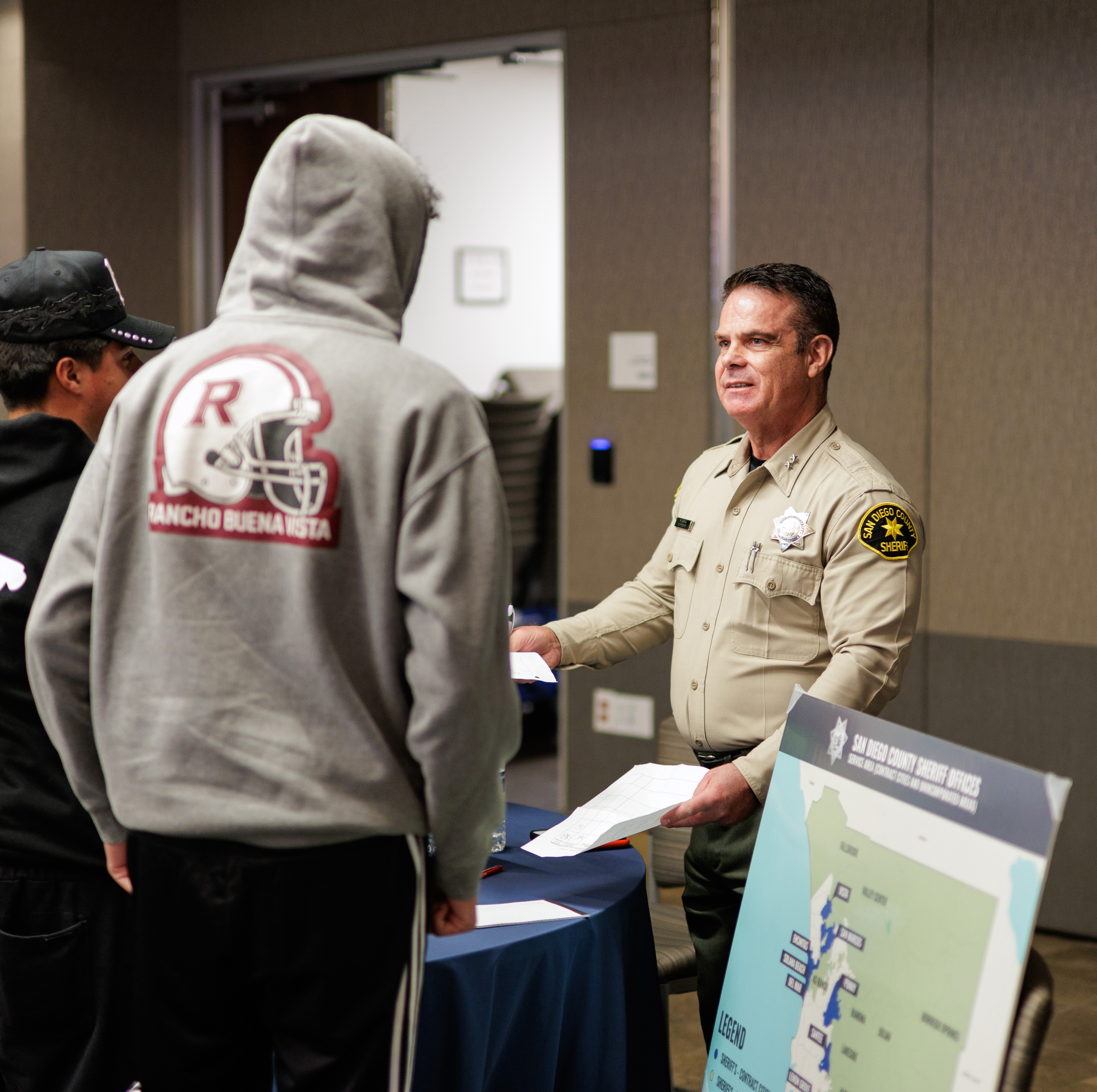 Assistant Sheriff Dustin Lopez talking to a person wearing a grey hoody with a table with a blue table cloth in the front discussing the Vista detention facility jail.