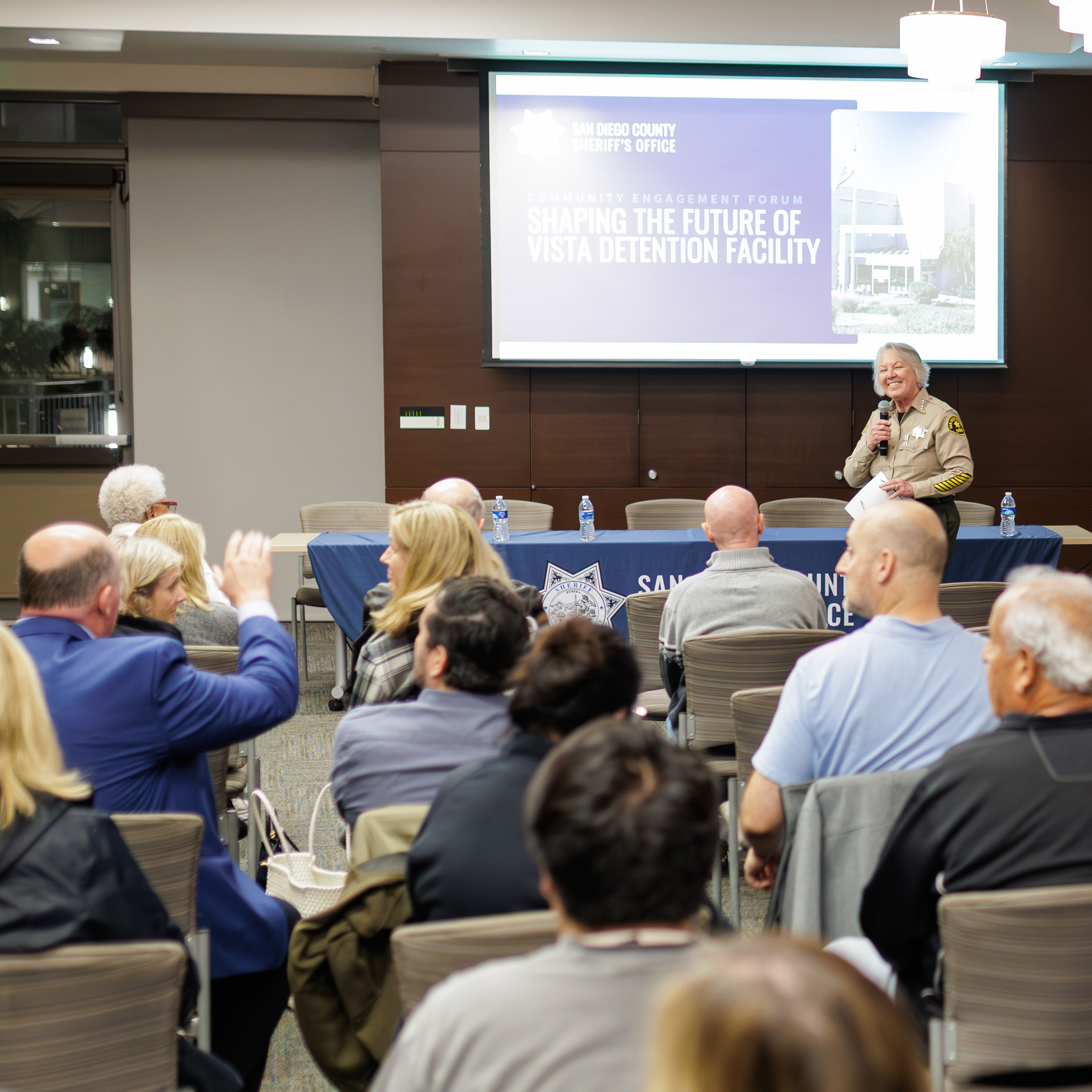 An indoor community meeting is underway with rows of seated attendees facing a speaker at a front table. The speaker, wearing a San Diego County Sheriff’s Office uniform, stands at a podium while addressing the audience. Behind the speaker is a large screen displaying a presentation titled ‘Shaping the Future of Vista Detention Facility.’ The room is well-lit with overhead lights, and several audience members appear to be engaging by raising hands or looking toward the front.