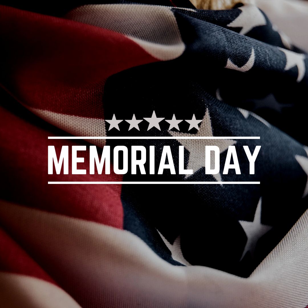 Close-up of a folded American flag showing red and white stripes and white stars on a dark blue field. Centered over the flag is white text that reads 'MEMORIAL DAY' with a row of five white stars above it and horizontal lines above and below the text.