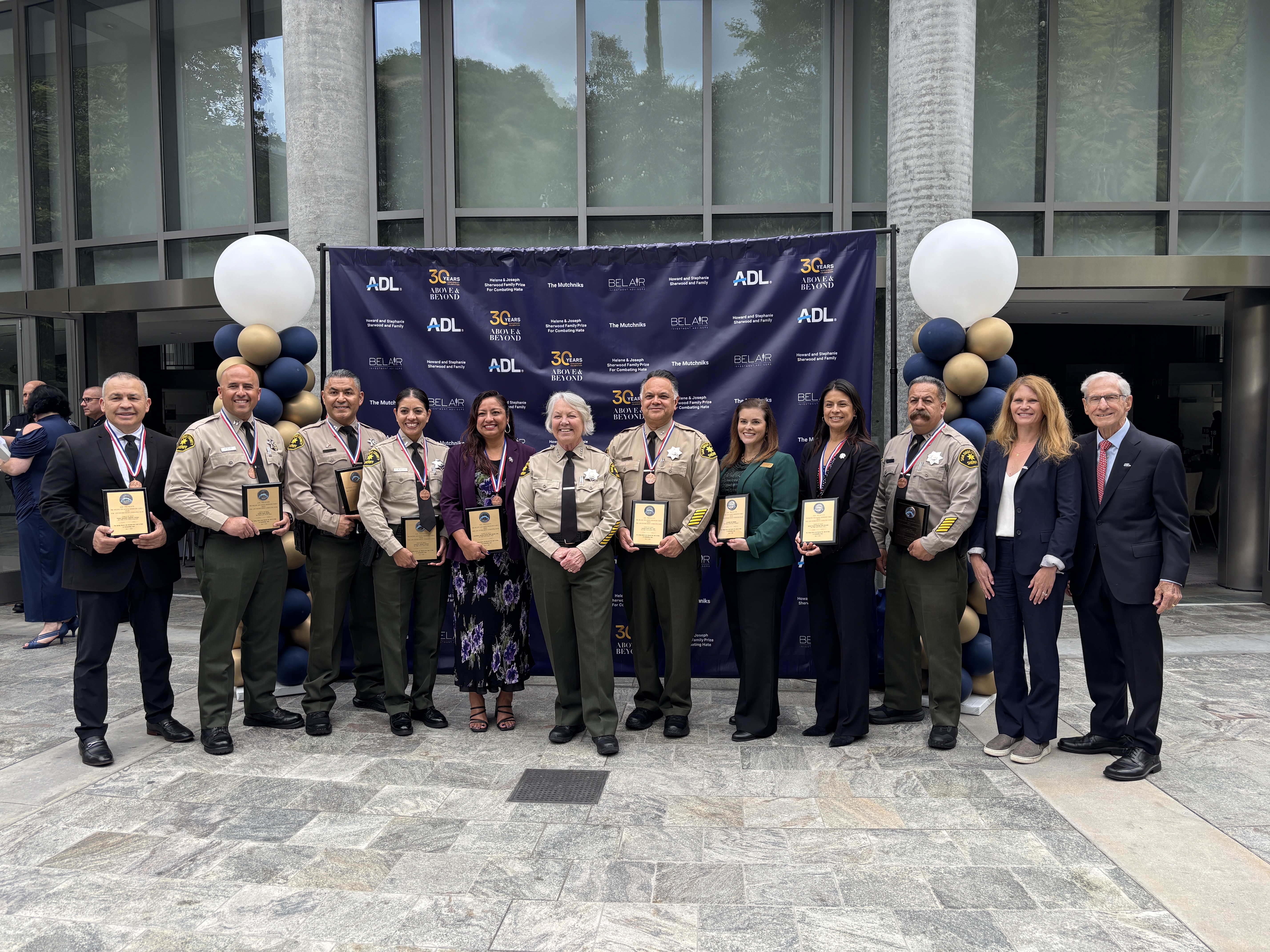 A group of uniformed Sheriff’s Department personnel and other attendees stand in a row holding award plaques in front of an ADL-branded backdrop decorated with balloon columns. The photo is taken outdoors in front of a modern building with large windows and stone pillars.