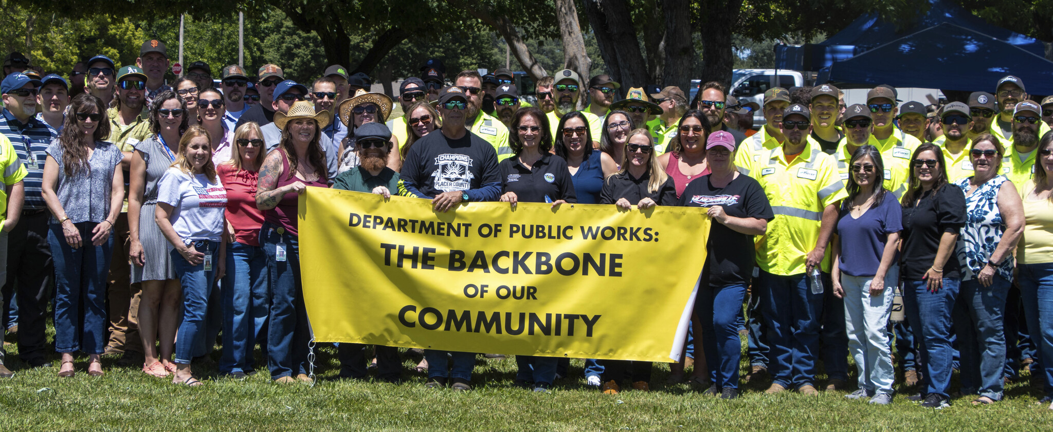 Crowd of community leaders and members of the Department of Public Works standing in green grass holding a yellow sign that reads "Department of Public Works: The Backbone of our Community"