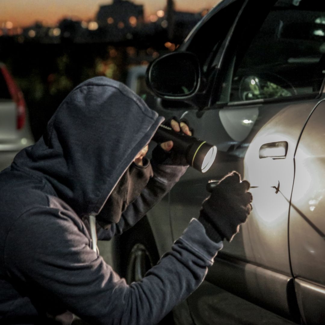 A person wearing a hooded sweatshirt is crouched beside a parked car at dusk, holding a flashlight and a tool up to the vehicle’s door. The scene is dimly lit, with city lights visible in the background.