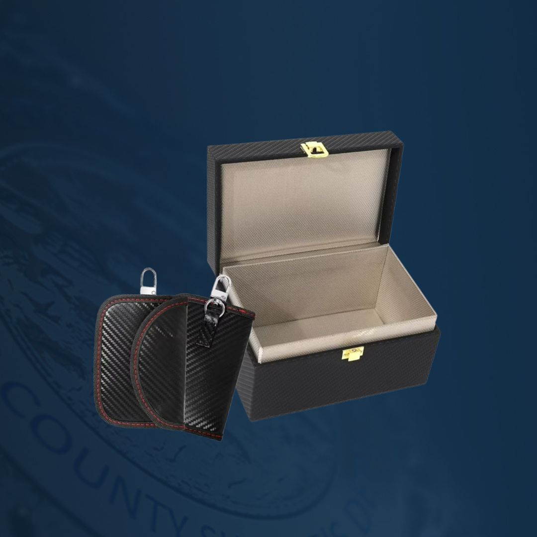 A small open lockbox with a light-colored interior is displayed against a dark blue background. Next to the lockbox are two textured signal‑blocking key pouches with metal keyring loops.