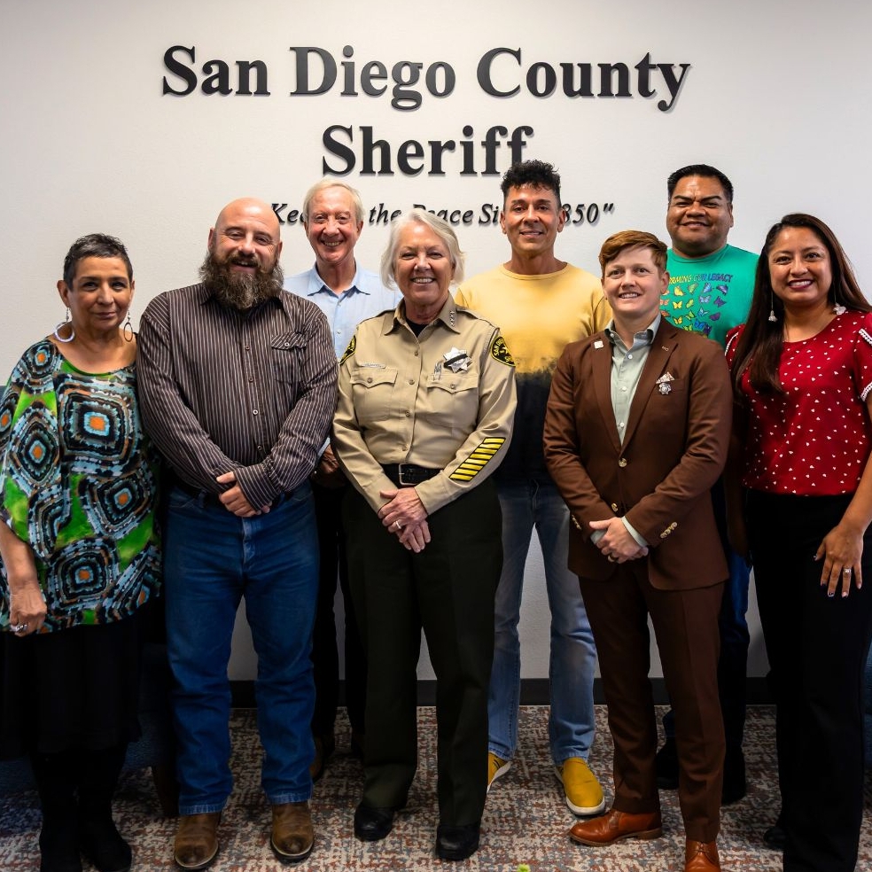 A group of nine people stand together in front of a wall with text that reads “San Diego County Sheriff.” The group includes individuals in uniforms and professional clothing, standing side by side and facing the camera. The background is a simple indoor setting with neutral walls and flooring.