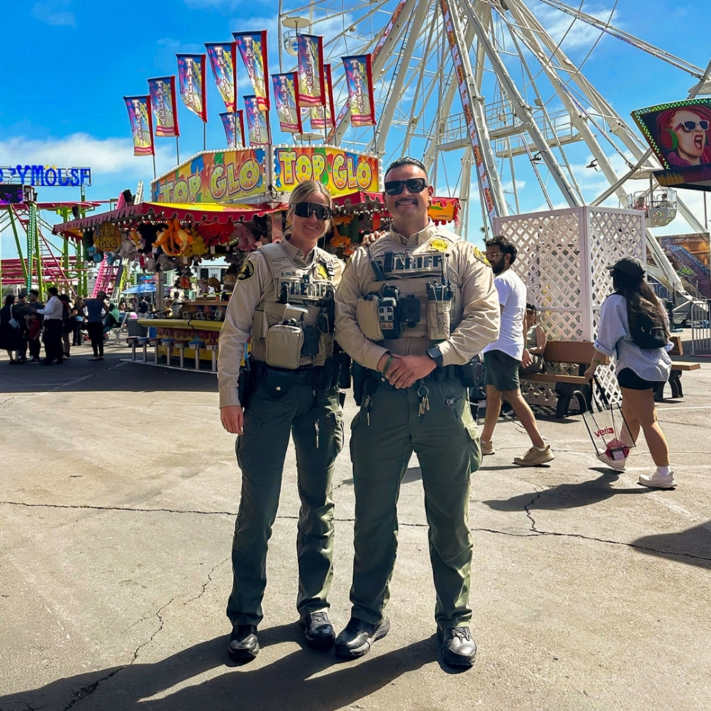 Two uniformed individuals stand side by side at an outdoor fairground, with a large Ferris wheel, colorful booths, and visitors walking in the background under a bright blue sky.