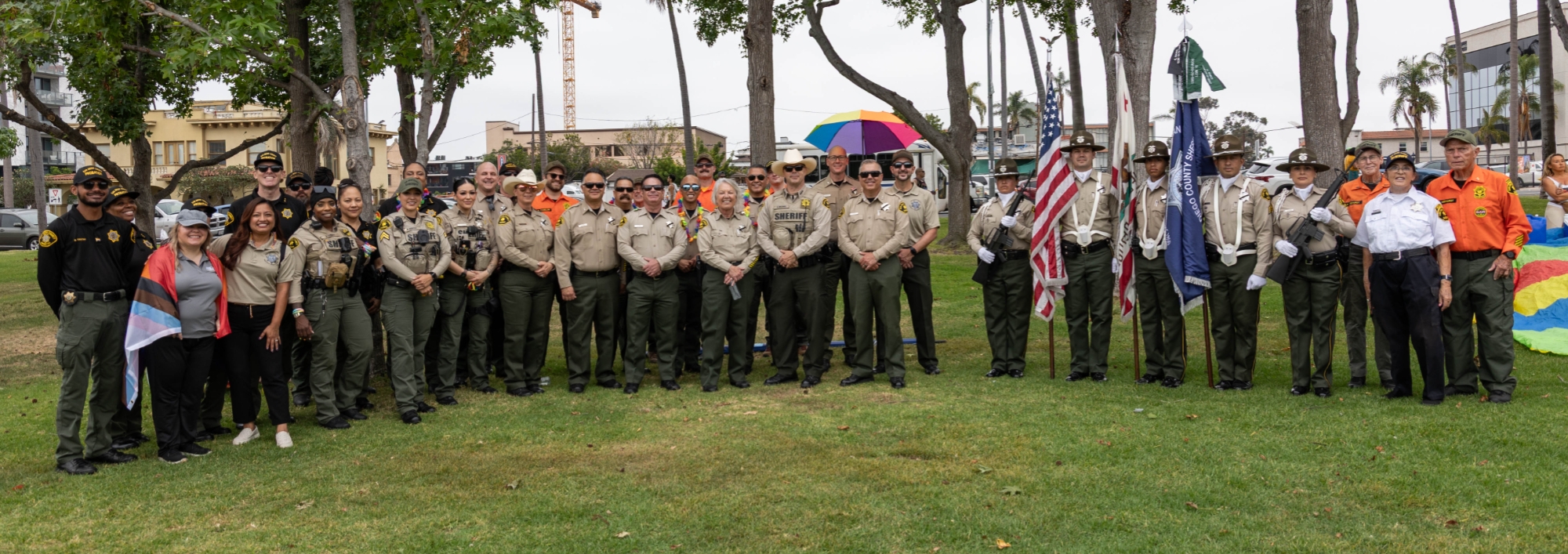A large group of uniformed and non‑uniformed individuals are gathered outdoors on a grassy area, standing in a wide formation. Trees and buildings are visible in the background, along with several colorful flags and umbrellas. The group faces the camera in an open park‑like setting.