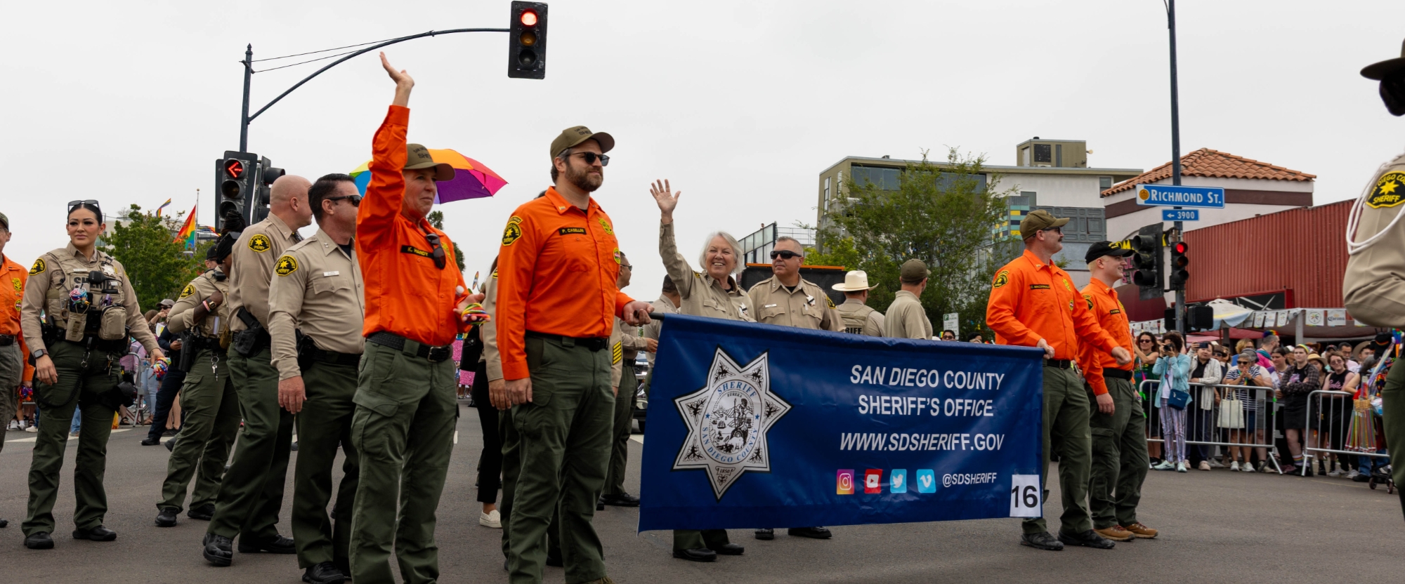 A group of uniformed and non‑uniformed individuals participate in a street parade, walking together and holding a large banner. Several people near the front of the group have one arm raised. A traffic light, buildings, and spectators lined along the street are visible in the background.