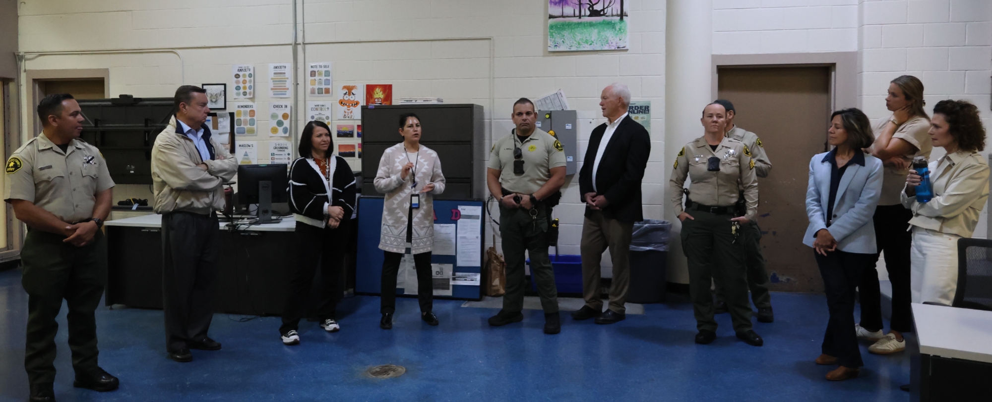 A group of people stand in a semi‑circle inside a room with blue flooring and light-colored walls. Several individuals wear uniforms, while others wear professional clothing. One person in the center is speaking to the group. Office equipment, cabinets, and a colorful poster are visible in the background.