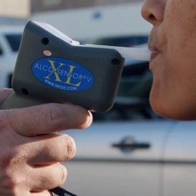 A close-up nighttime scene showing a handheld alcohol-screening device held by a uniformed deputy near an individual who is standing beside others. A red canopy with partially visible text is overhead, and the background includes blurred lights and figures, suggesting an active enforcement area. Let me know if you’d like a shorter or more formal version!