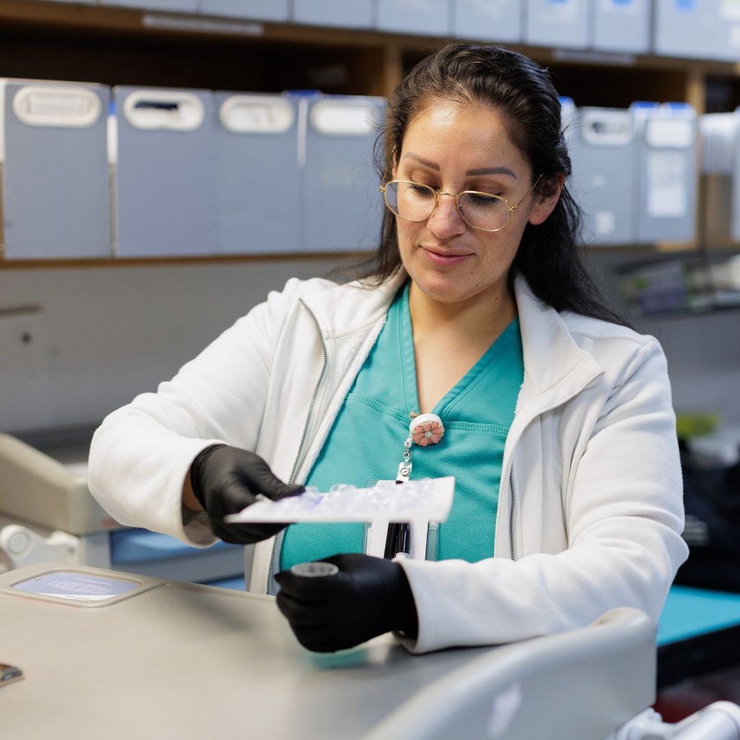 A nurse with lab jacket and medicine with glasses and turquoise shirt with files behind her.
