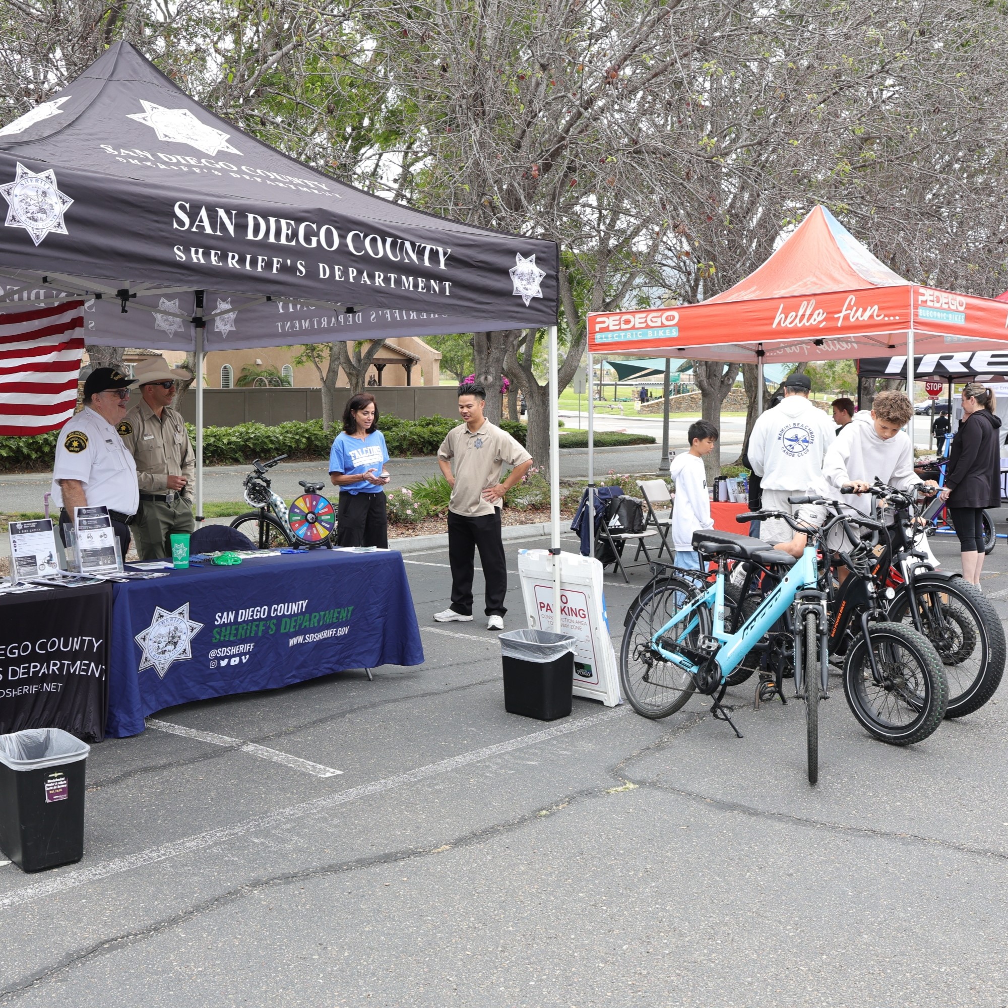 A ceremonial scene at an outdoor law enforcement memorial, where two uniformed honor guard members stand before a large engraved memorial wall. One individual raises a hand in a formal salute. A wreath decorated with red, white, and blue flowers is positioned beside the wall. In the foreground, a badge‑shaped emblem displays the text “San Diego County Law Enforcement Memorial Foundation.” An overlay of a dark blue American flag with stars frames the composition.