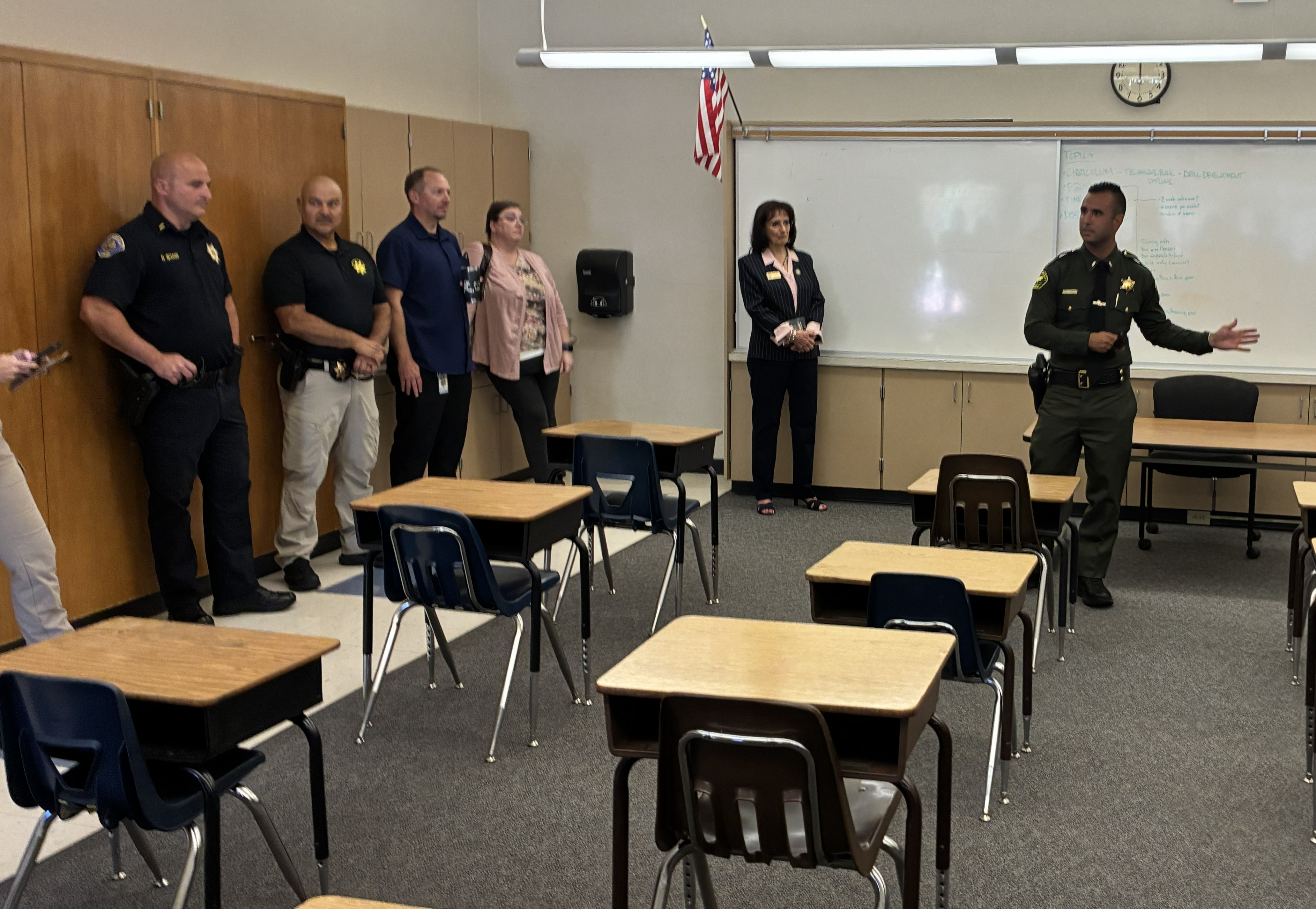 Supervisor Jones standing under an American flag in front of a white board in a room set up like an elementary school classroom with peace officers and stakeholders listening to Placer County Sheriff's Lt. Michael Cuevas as he guides their tour through the new facility.