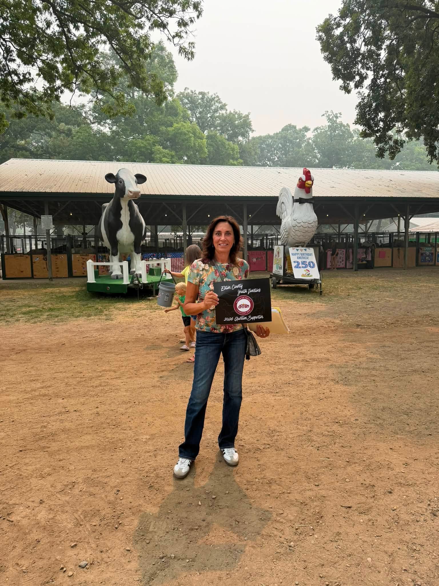 State Representative Angela Witwer State Representative Angela Witwer at the Eaton County fair holding a banner that reads "Eaton County youth livestock 2026 auction supporter" while standing in front of statues of a cow and a chicken.