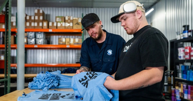 Two men examining a set of light blue shirts with screen printed on black logos.