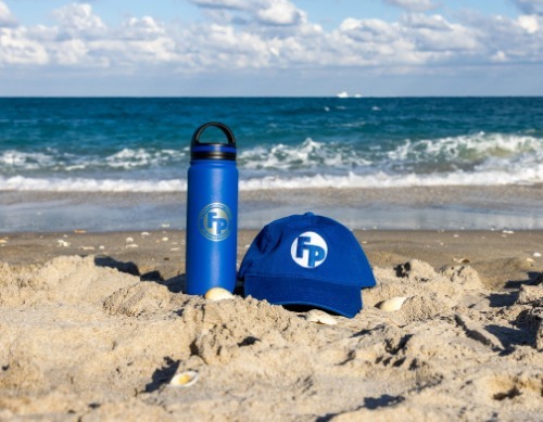 A blue water bottle with a screen printer on Fully Promoted Logo on the sand in a beach next to a blue hat with  an embroirdered Fully Promoted logo.