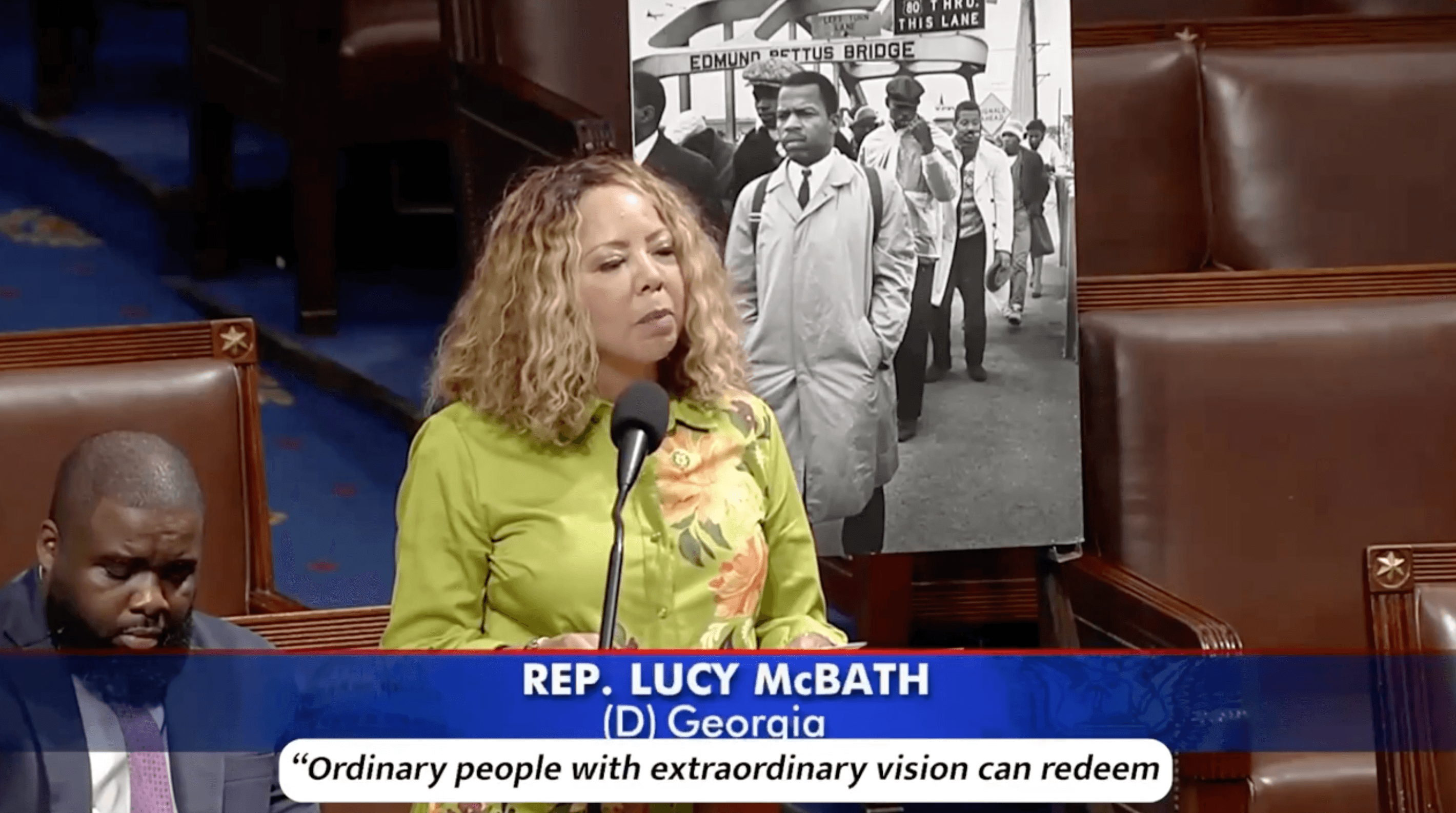 Rep. McBath speaking from the floor of the U.S. House of Representatives in front of a black and white photo of John Lewis walking across the Edmund Pettus Bridge.