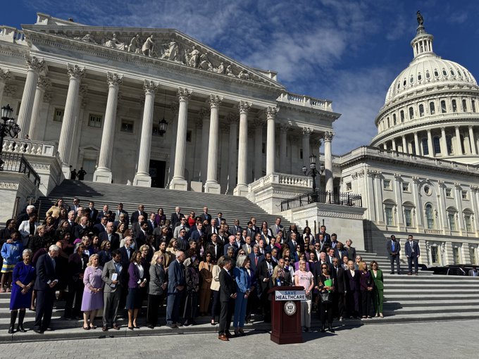 Members of the Democratic Caucus stand on the steps of the U.S. House behind a podium which bears a sign reading "Save Healthcare".