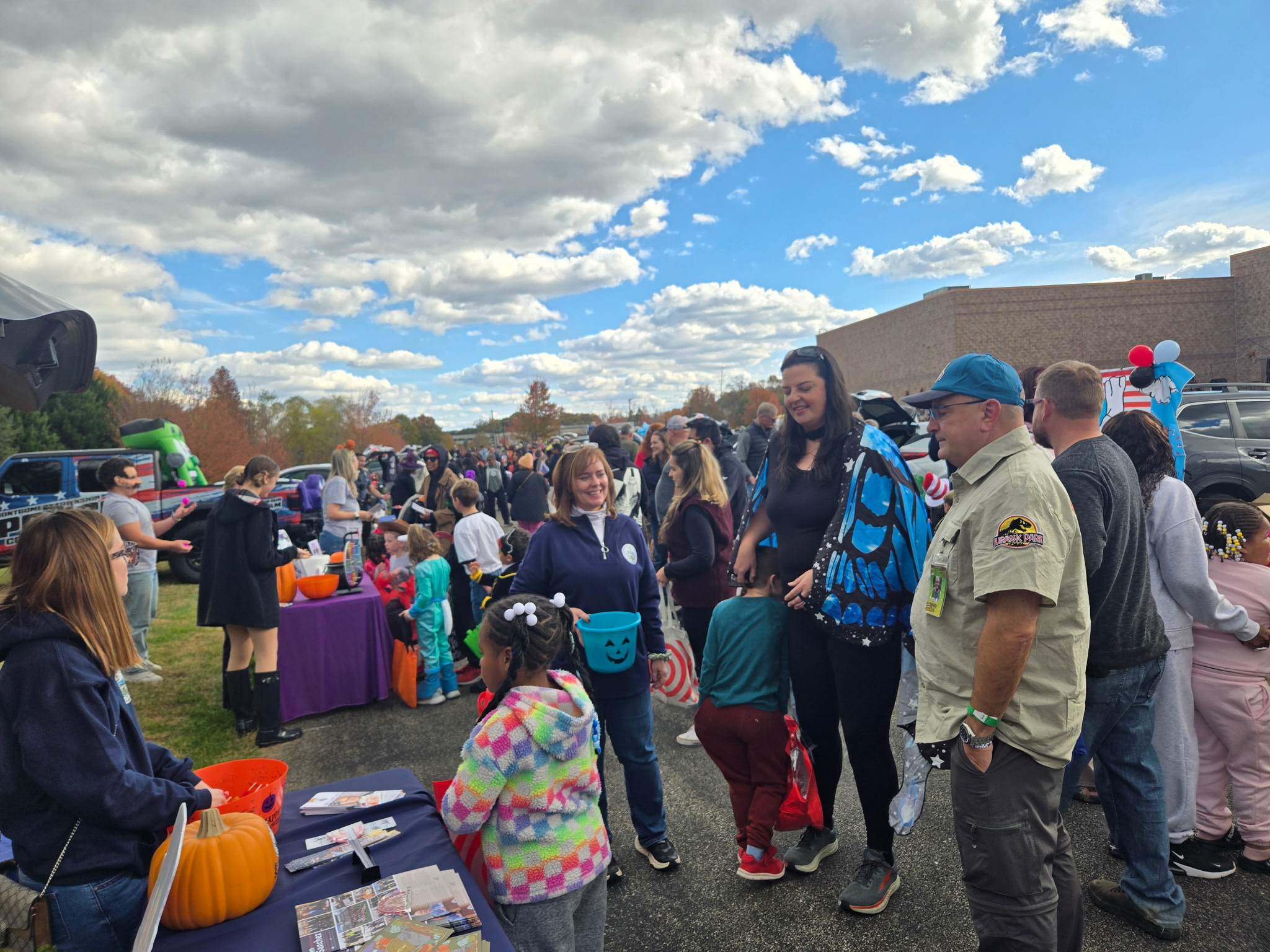A photo of a large crowd of people walking around the trunk or treat resource fair with Rep Sanchez, Rep Hanbidge and staff at the table in the foreground with children.
