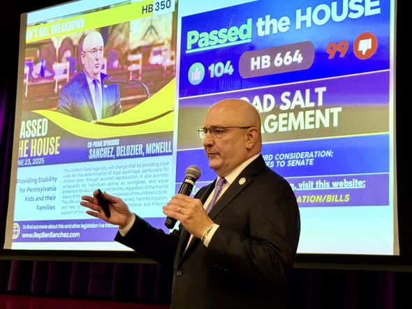 Rep Sanchez speaking into a microphone with an image of two bills he passed recently projected on a screen behind him.